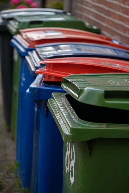 Five large wheeled rubbish bins made of dark grey plastic with yellow lids are lined up side by side against a plain, light grey wall. Each bin has a white oval label with printed information and a un