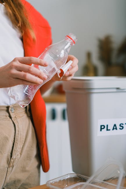 A waste collection worker wearing a high-visibility yellow vest and green trousers operates the controls of a white refuse collection truck parked on a city street during evening hours. The back of th